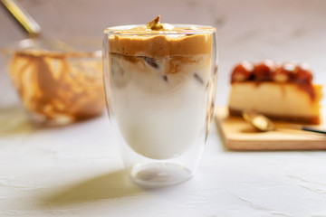Dalgona Coffee; fluffy whipped coffee on milk with whisking bowl of coffee and caramel macadamia cheesecake on wooden tray.