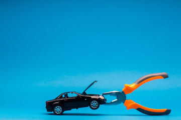 A black car figurine caught in a pair of pliers, on blue background.