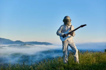 Tuinposter Astronaut wearing white space suit and helmet playing white guitar, standing on sunny green mountain glade in summer under blue sky, morning fog rising up from the valley behind him. © anatoliy_gleb