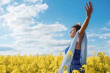 Happy woman wearing blue top and jeans  raised her hands high against blu sky and yellow rapeseed fields