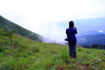 Naklejka premium Asian hiking or tourist woman in dark blue sweater with camera standing on green grass field with mountain view and mist background and copy space. Beautiful in nature and Landscape