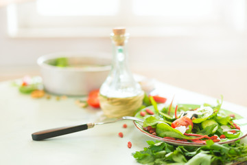Fototapeta na wymiar Healthy cuisine. Full bowl of fresh salad served on light kitchen table with olives oil and fork at sunny window background. Diet or vegetarian food concept. Home food eating