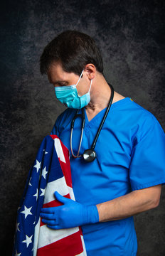Sad/pensive male doctor in blue hospital scrubs with face mask and stethoscope, nursing the Stars & Stripes American flag close to his chest, and standing against a dark studio background.