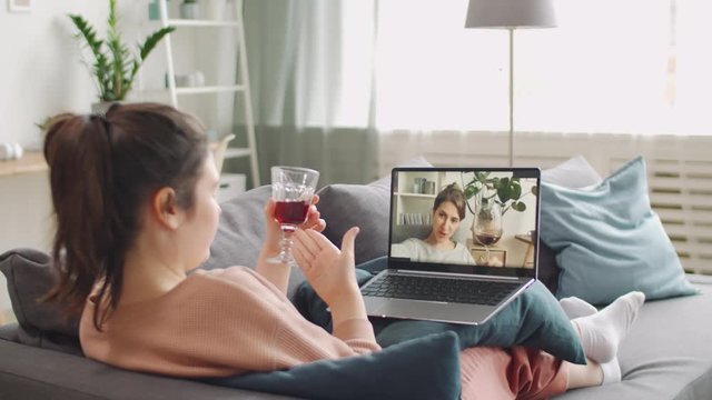 Young woman sitting on couch with laptop, chatting with female friend on video call, giving a toast and drinking red wine while staying at home on quarantine
