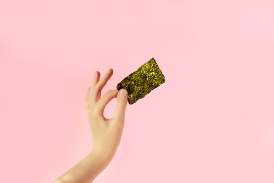 Female hand with crispy seaweed on the pink background with copy space. Dry nori sheets korean or japanese roasted food texture. Flat lay.