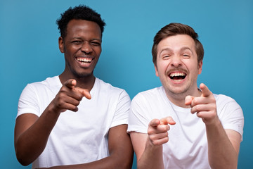 Two man african american and caucasian pointing at camera with index finger and laughing out loud