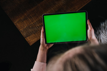 Young woman using black tablet device with green screen. Woman holding tablet, scrolling pages while sitting on the couch in the living room. Chroma key. View over shoulder. Close up