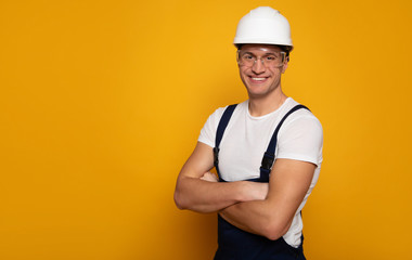 Friendly builder. Close-up photo of a young man in a working clothes, who is wearing a helmet and transparent glasses, while standing with his arms folded.