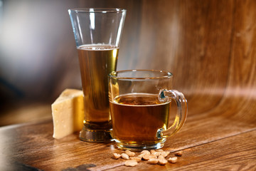 A glass and a mug of tasty beer stand on a brown background with cheese and nuts. Fragrant beer lit by beautiful light stimulates appetite.