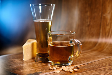 A glass and a mug of tasty beer stand on a brown background with cheese and nuts. Fragrant beer lit by beautiful light stimulates appetite.