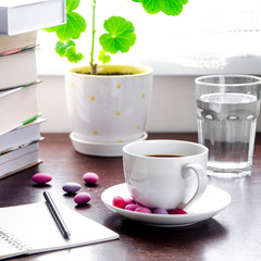 A white cup of coffee with multi-colored sweets stands on a wooden table, next to a stack of books and a notebook against