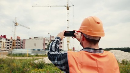 A man in an orange vest and an orange helmet is filming a construction video on a mobile phone.