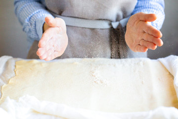 Obraz na płótnie Canvas Hands in flour. A home-baker in an apron rolls the dough and sprinkles it with flour. Flour scatters at the hands. Girl sprinkles flour with dough. Cooking pasta from the dough. Hand-wrapping flour