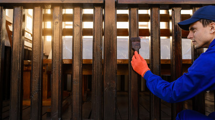 Contractor worker in blue overalls and protective gloves paints a fence