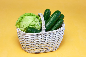 Small white basket with fresh green cabbage and cucumbers on yellow background. Vegetables in basket close up.