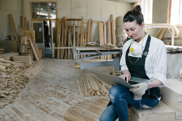 Confident woman working as carpenter in her own woodshop. She using a laptop and writes notes while being in her workspace. Small business concept.