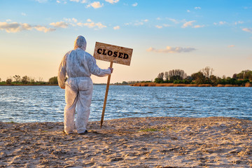 Male wearing protective suit stands with his back with warning sign in hand with inscription closed along sandy beach of river. Contaminated water, quarantine, virus outbreak, crime scene concept