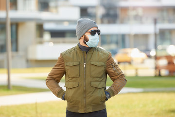 A man in a medical face mask to avoid the spread coronavirus walking in the cozy street. A fellow dropping his hands in pockets wears a cap, sunglasses, and a face mask against COVID 19.