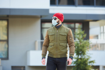 A man in a medical face mask to avoid the spread coronavirus holding his smartphone in a cozy street. A guy trekking wears a red cap, sunglasses, and a face mask against COVID 19.