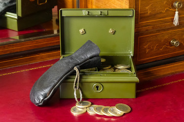 Old open green metal petty cash tin, with a black leather coin bag. Coins in tray and on the desktop  Sitting on a tooled red skiver of a Victorian  writing bureau. Reflection in mirror. England.  