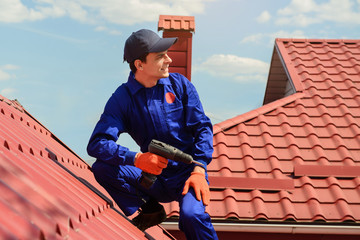 Young happy man contractor worker in blue overalls is repairing a red roof with electric screw...