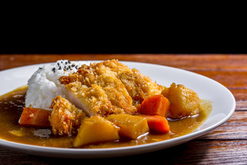 Japanese curry with rice, black sesame, carrot, potato and deep fries chicken in white dish on wooden table.