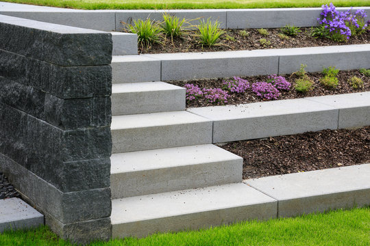 Moderne terrassenförmige Gestaltung eines Garten  Hanggrundstücks: Treppe und Stufen aus Betonsteinen mit Gräsern und lila Blumen geschützt mit Mulch und begrenzt von einer grauen Naturstein Mauer 