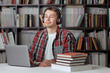 Happy guy listens to a remote audio lesson in the library with wireless headphones and prints a report on a laptop.