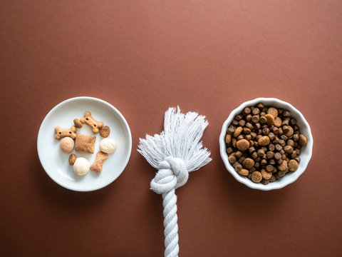 Hunde Trockenfutter und Leckerlies in zwei wei&szlig;en Sch&uuml;sseln und Hundespielzeug auf einem braunen Hintergrund, Flat lay, Tierthema