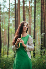 Woman in a green medieval dress plays the wooden flute while standing in a grove against the background of pines. Girl in a gloomy forest plays music on a handmade pipe. Young witch in a gloomy forest © Михаил Гута
