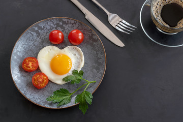 Breakfast with fried eggs, tomatoes and a cup of coffee. Fork and knife on a black background. Copy space.
