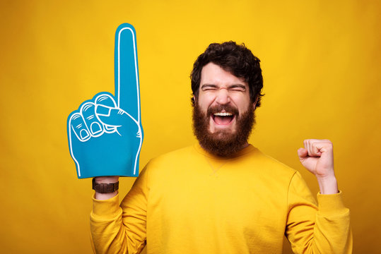 Excited young man is wearing a foam fan glove while making winner gesture on yellow background.
