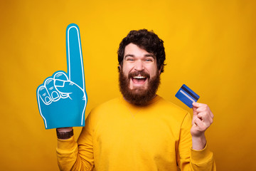 Excited bearded man is holding a credit or debit card and wearing a foam fan glove on yellow background.