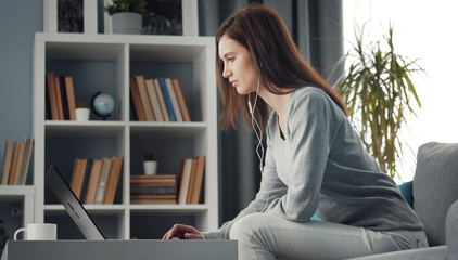 Side view of young woman with earphones sitting on couch in front of laptop on coffee table, low angle