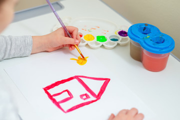 Child is drawing the sun near drawn red house with watercolors on the white sheet of paper.