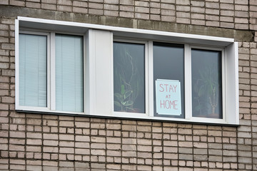 Banner with inscription stay at home, stands behind the glass on window of brick house.