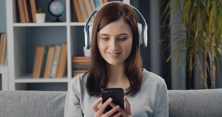Headshot of glad young female using smartphone and wireless headphones to divert herself at home