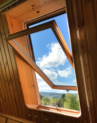 opened roof  window with wooden frame on mansard floor with beautiful view of blue sky white clouds and green forest outside, luxury wooden house