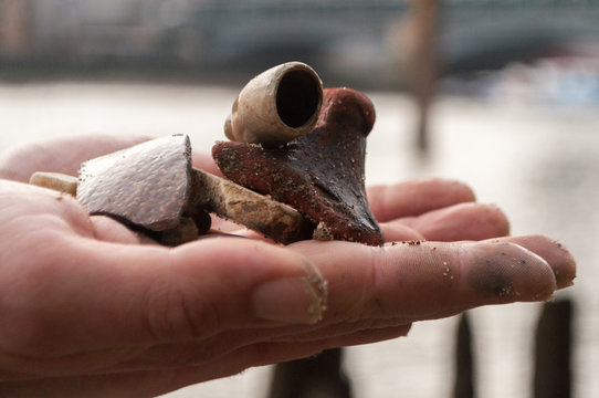 Mudlark Findings: A man's hand holds an old broken clay pipe bowl and other pieces of the past he has found while mudlarking on the River Thames