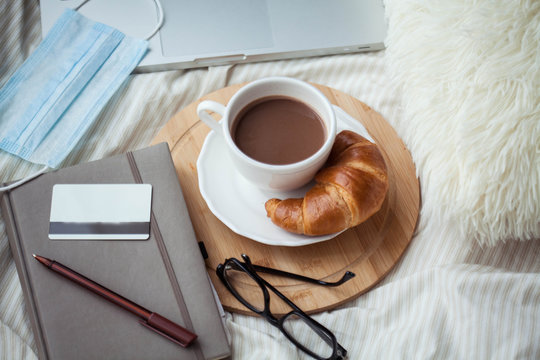 A cup of hot cocoa, a croissant, a wooden stand, a medical mask, a notebook, a laptop, glasses, a bank card on the bed. Quarantine time. Home Office. Online shopping.