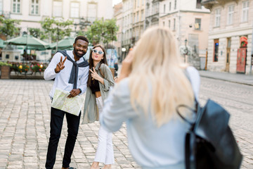 Excited cheerful mix raced couple, African man and Caucasian girl, enjoying walking outdoors, in the center of ancient European city, while their pretty female friend is making photo of them