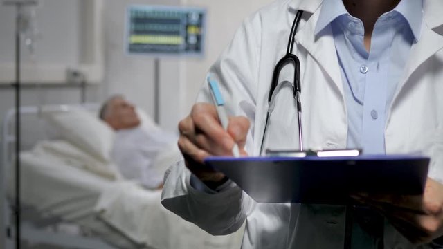 close up of doctor writing report diagnosis of patient lying on the hospital bed in the background
