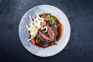Traditionelle deutsche geschmorte Ochsenbacken in brauner Rotwein Sauce mit Nudeln und Karotten angeboten als Draufsicht auf einem Modern Design Teller auf einem rustikalen Board mit Textfreiraum