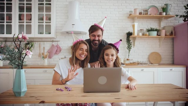 Happy family with a daughter waving hands and celebrating birthday in kitchen using laptop for a video call during online birthday party