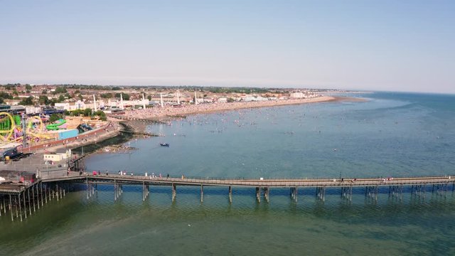 Drone or aerial footage of Southend pier on a busy sunny bank holiday. The beach and sea are full of people and families playing and swimming close to a funfair with a rollercoaster