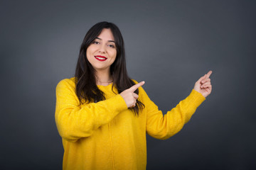 Pretty young brunette woman with positive expression, dressed casually, indicates with fore finger...