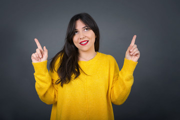 Horizontal shot of successful friendly looking young dark haired woman with bright make up exclaiming excitedly, pointing both index fingers up, indicating something shocking on blank copy space wall.