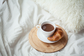 A cup of hot cocoa, a croissant, a wooden stand on the bed. Quarantine time. Homeliness.