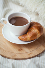 A cup of hot cocoa, a croissant, a wooden stand on the bed. Quarantine time. Homeliness.