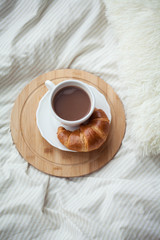 A cup of hot cocoa, a croissant, a wooden stand on the bed. Quarantine time. Homeliness.